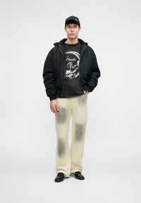Schwarze Pufferjacke über einem grafischen Sweatshirt, cremefarbene, weite Hose mit dunkleren Akzenten und schwarze Schuhe. Eine schlichte Mütze rundet den Look ab.