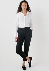 Wit overhemd met knopen en lange mouwen, zwarte op maat gemaakte broek, en zwarte loafers met een gouden kettingdetail. Gladde stof, strakke pasvorm.