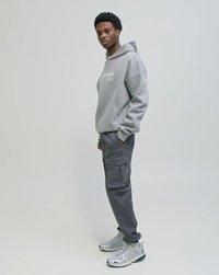 Junger Mann steht seitlich, trägt einen grauen Hoodie, dunkelgraue Cargohosen und silberne Turnschuhe vor einem schlichten hellen Hintergrund.