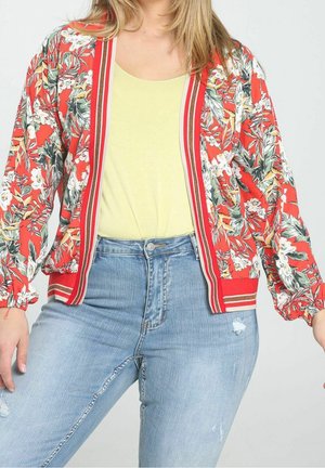 Femme portant une veste bomber rouge à fleurs, un top jaune pâle et un jean bleu clair taille haute déchiré, sur un fond uni.