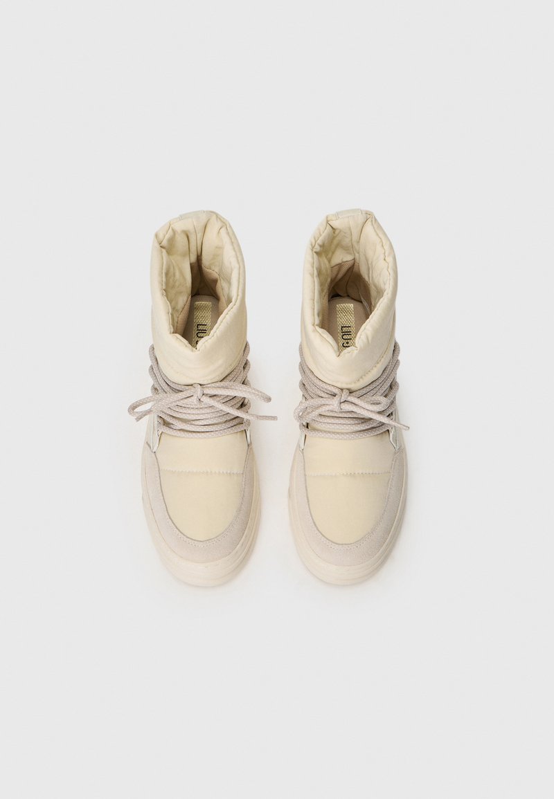 Zapatos altos beige con parte superior acolchada, tela texturizada y cordones en tonalidades similares. La suela de goma está ligeramente elevada y tiene un acabado suave.