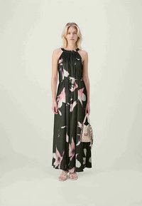 Mulher vestindo um vestido maxi preto sem mangas com estampa floral rosa e branca, segurando uma bolsa bege e usando sandálias brancas com tiras.