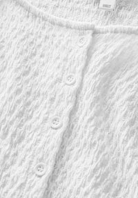 Cardigan blanc en tissu texturé avec un motif ondulé, doté d'un col rond et de cinq boutons blancs à l'avant.