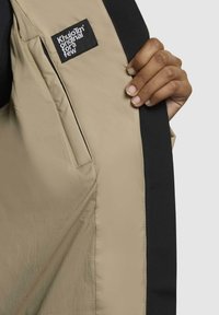 Fodera interna khaki di un giubbotto, con un bordo esterno nero. Un'etichetta legge "Khujon Original per pochi." Mano che tiene il tessuto.