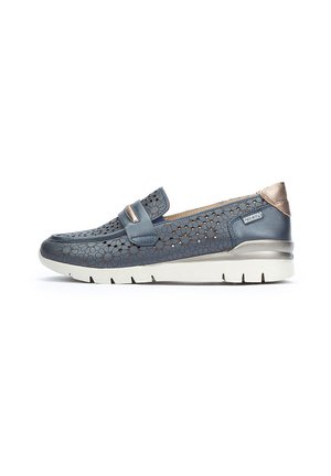Zapato azul de piel perforada tipo slip-on con acentos plateados, suela blanca acolchada y un diseño plano que presenta una tira ancha sobre el empeine.
