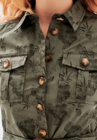 Camicia verde oliva con motivo floreale, dotata di due tasche sul petto, bottoni marroni e colletto. Il tessuto ha una consistenza liscia.