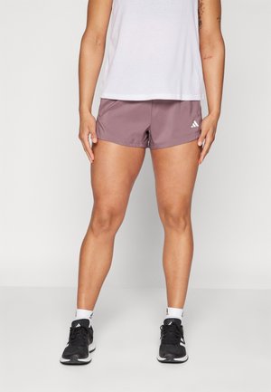 Person wearing mauve Adidas athletic shorts, white tank top, white Adidas socks, and black Adidas running shoes standing on a gray floor.