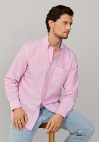 Man met donker haar, gekleed in een lichtroze overhemd met knopen en lichtblauwe spijkerbroek, zittend op een houten krukje, kijkt naar rechts.