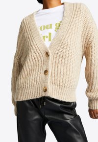 Gruby kardigan w jasnym beżu z dekoltem w serek, czterema drewnianymi guzikami, prążkowaną teksturą i luźnym krojem. Połączony z czarnymi skórzanymi spodniami.