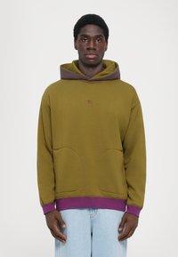 Felpa verde oliva con cappuccio marrone, caratterizzata da un orlo viola e due tasche frontali. Texture morbida, design minimalista e dettagli con logo ricamato.