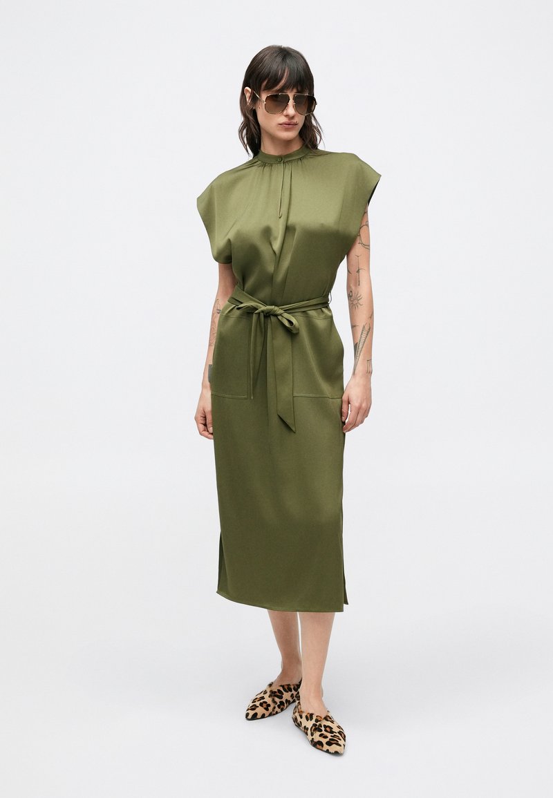Olive green, knee-length dress with cap sleeves, a tie waist, and side pockets. Worn with leopard print flats and sunglasses.