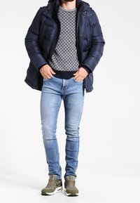 Doudoune marine avec capuche, pull gris à motifs, jean bleu slim et baskets vert olive avec semelles blanches.