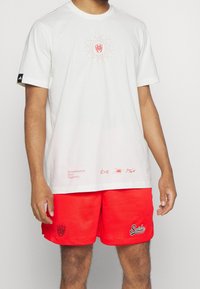 Valkoinen puuvillateeppari, jossa on keskeinen grafiikka ja teksti, yhdistettynä punaisiin verkkohousuihin, joissa on logo ja tekstityksellä varustettu yksityiskohta.