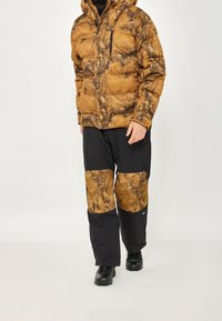 Braune und schwarze isolierte Jacke mit einem marmorierten Muster, kombiniert mit schwarzen Hosen, die braune Akzente und mehrere Taschen aufweisen.