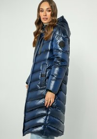 Parka blu navy con finitura lucida, dotata di cappuccio, accenti neri e motif a righe orizzontali. Include tasche laterali e coulisse.