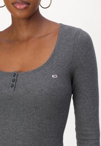 Frau trägt ein graues, geripptes Langarmoberteil mit drei schwarzen Knöpfen und einem kleinen rot-weißen-blauen Logo auf der Brust.