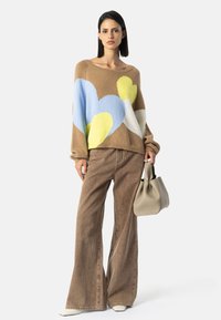 Kvinne med lys brun genser med store blå, gule og hvite hjerter, brune vide bukser, hvite sko, og holder en lysebeige håndveske.