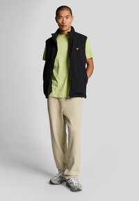 Gilet nero senza maniche sopra una maglietta verde chiaro, pantaloni beige e scarpe sportive grigie con dettagli bianchi, in piedi davanti a uno sfondo grigio chiaro.