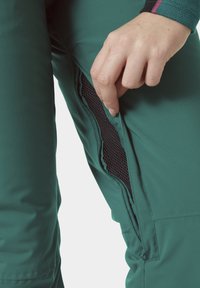 Groene geïsoleerde broek met een gaasventilatie-detail, voorzien van een zijrits en een gladde buitenkant. De hand wordt getoond terwijl deze de rits naar beneden trekt.