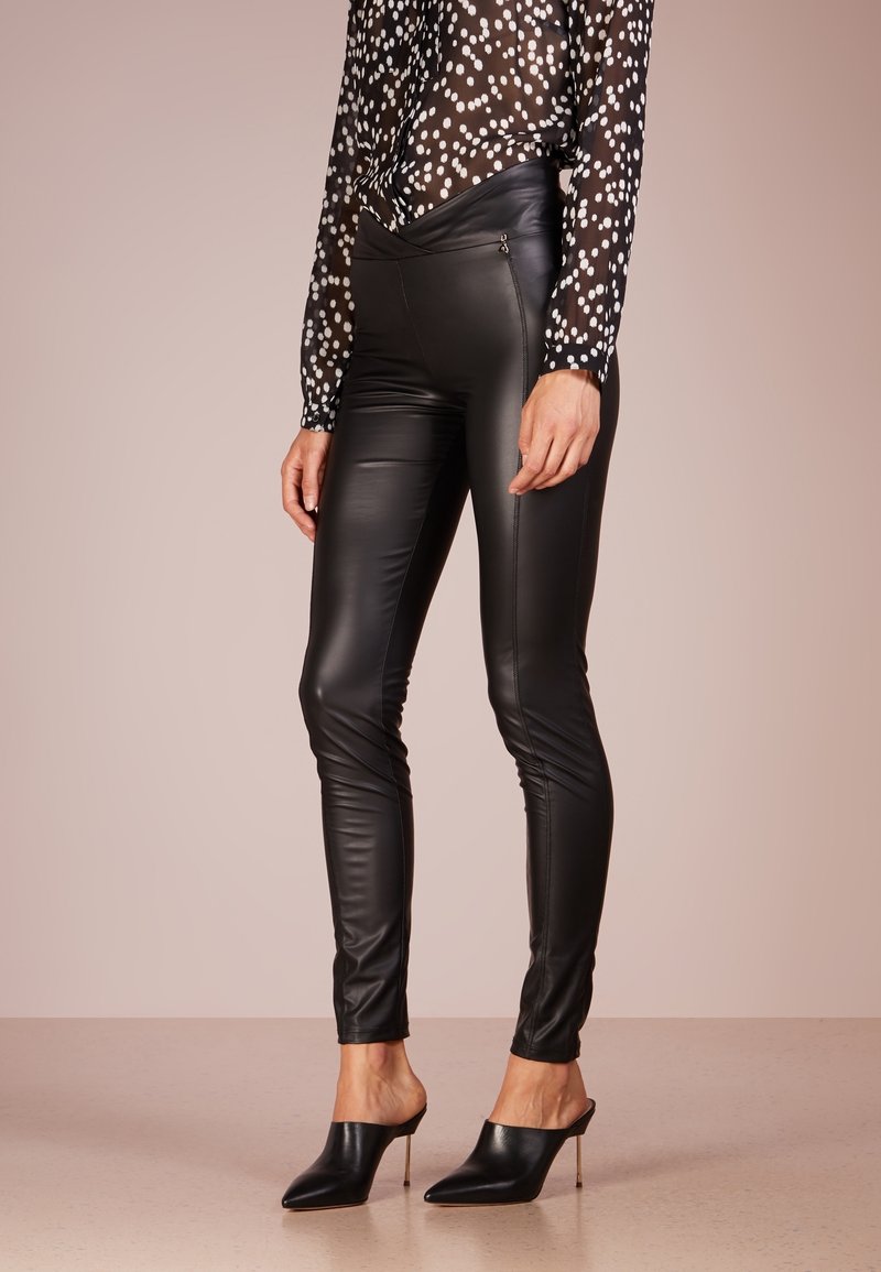 Black faux leather leggings with a slim fit, featuring subtle side zippers. Paired with a sheer black blouse dotted in white.