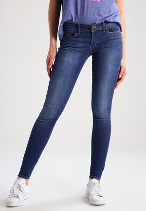 Personne portant un jean bleu ajusté, des baskets blanches et un haut violet partiellement visible, debout, jambes écartées, sur un fond uni.