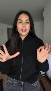 Fiatal nő hosszú sötét hajjal, fekete cipzáras pulóvert és farmert viselve, kézmozdulatokat téve, miközben bent beszél.