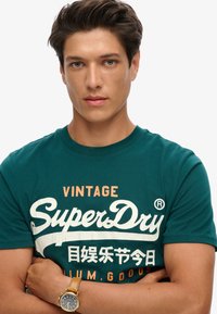 Camiseta de manga corta color verde azulado con el logo "Vintage SuperDry" en blanco y naranja. Tejido suave; sencillo cuello redondo; combinada con un reloj marrón.