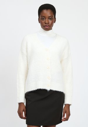 Witte gebreide cardigan met V-hals, vijf grote knopen, geribde manchetten en een iets kortere zoom, gedragen over een witte coltrui en een zwarte rok.