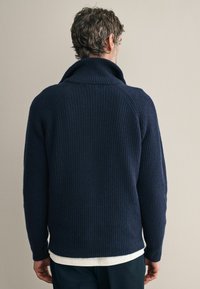 Pulover din tricot cu fir în dungi, de culoare bleumarin, cu guler înalt, prezentând un finisaj texturat, mâneci raglan și o croială relaxată. Cămașă albă vizibilă dedesubt.