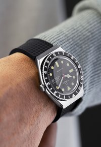 Quartz wristwatch with a black dial, luminous hour markers, stainless steel case, and black patterned silicone strap. Date display at 3 o'clock.