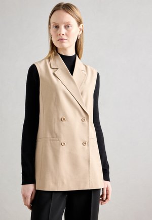 Beige double-breasted vest with notched lapels, two buttons, and side pockets, worn over a black long-sleeve top. Smooth fabric texture.