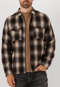 Brun og gråternet flanneljakke med to brystlommer, lynlås foran og kontrastfarvet ribstrikket sweater under. Blød tekstur, afslappet design.