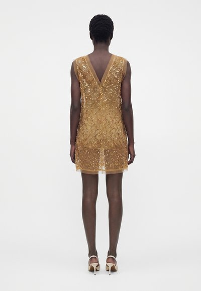 Femme portant une robe courte dorée sans manches avec broderie en sequins et un dos en V profond, debout les bras le long du corps et chaussée de talons hauts beige.