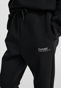 Person iført sorte joggingbukser med snøre i taljen og lille hvid hummel-logo på det øverste lår, hænder i lommerne.
