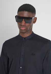 Camicia nera a maniche lunghe con colletto alla coreana, caratterizzata da dettagli di cucitura discreti e un logo ricamato sul petto. Occhiali da sole neri con design angolare.