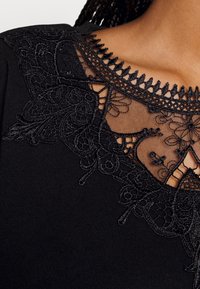Top nero con dettagli in pizzo nero intricati sullo scollo, che presenta motivi floreali e bordi a scallop su un tessuto morbido.