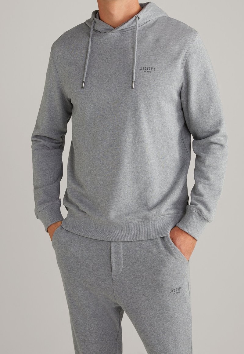 Sudadera gris y pantalones a juego hechos de un tejido suave. Presenta un bolsillo tipo canguro, capucha con cordón y una discreta marca en el pecho.