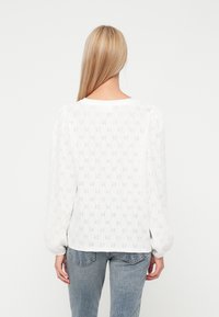 Witte lange mouwen shirt met een oogjespatroon, ronde halslijn en geplooide mouwen. Gecombineerd met een medium-wash spijkerbroek.