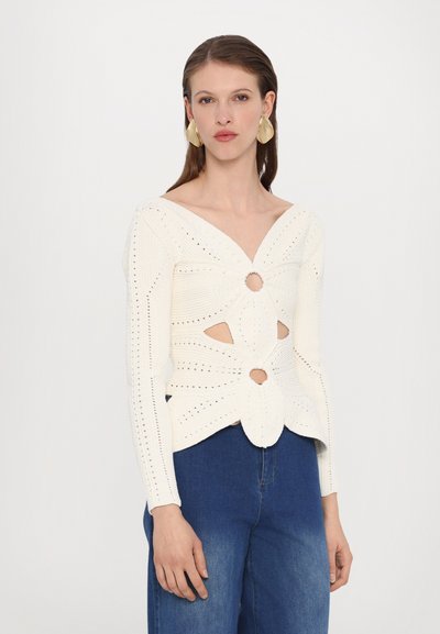 Knit, cream-colored top with V-neck, cut-out details, and textured patterns. Long sleeves; paired with high-waisted blue denim pants.