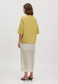 Gelbes Strickoberteil mit kurzen Ärmeln, kombiniert mit einem transparenten weißen Maxirock und schwarzen Absatzsandalen, von hinten betrachtet.