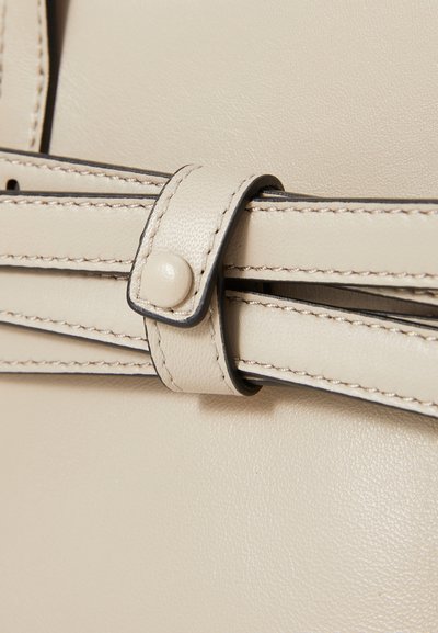Gros plan sur un sac à main en cuir beige avec une bandoulière cousue, un bouton circulaire et des détails de bord noir contrastés. Texture lisse.