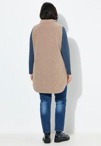 Gilet lungo beige trapuntato con colletto alto, realizzato in tessuto testurizzato, indossato sopra un top blu a maniche lunghe e abbinato a jeans e scarpe nere.