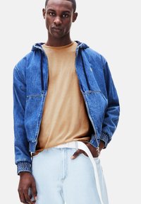 Giacca di jeans con cappuccio, di colore blu, polsini elasticizzati e zip frontale. T-shirt beige sotto, abbinata a jeans azzurri e una cintura bianca.