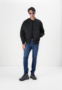 Chaqueta bomber negra con puños acanalados, camiseta negra ajustada, jeans azules y zapatillas negras. El modelo lleva gafas de sol blancas. Fondo simple.