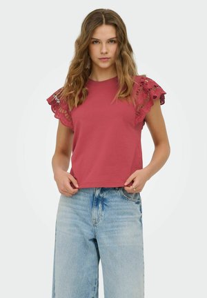 Jeune femme aux cheveux ondulés portant un haut rouge sans manches avec des détails en dentelle et un jean taille haute bleu clair, posant sur un fond uni.