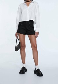 Žena v bílé zkrácené košilce s knoflíky, černých šortkách s vysokým pasem a knoflíky, bílých ponožkách, masivních černých botách a držící malé černé kabelky.