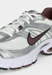 Λευκό και ασημί παπούτσι τρεξίματος Nike με καφέ λεπτομέρειες, διχτυωτό επάνω μέρος, μαλακή σόλα και το εμβληματικό καφέ λογότυπο Nike Swoosh στο πλάι.