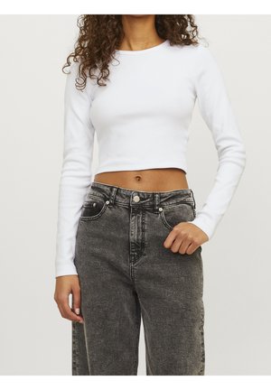 Persoon die een wit cropped shirt met lange mouwen en een hoog getailleerde, vervaagde zwarte spijkerbroek draagt, staand met één hand bij de zak.