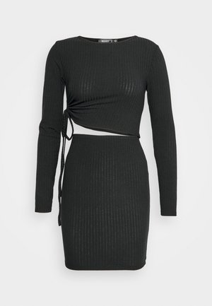 Robe mini noire en maille côtelée à manches longues, avec détail noué sur le côté et découpe asymétrique à la taille.