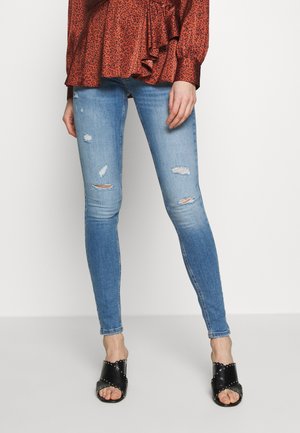 Vaqueros ajustados azules con detalles desgastados y rotos, combinados con sandalias negras deslizantes con acentos de tachuelas. La parte superior es roja con estampado de leopardo.